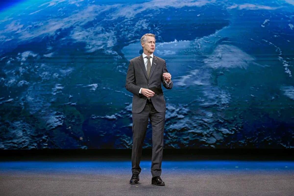 A man in a dark suit, white shirt, and dark tie is standing on stage delivering a keynote speech. He is making a gesture with his right hand while speaking. The background features a large screen displaying an image of Earth from space, with blue oceans and white cloud formations visible.