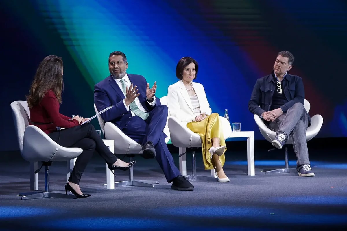A panel discussion at MWC25 featuring four professionals seated on a stage with a modern, abstract blue and green background. One panelist, a man in a blue suit, is actively speaking and gesturing with his hands, while the others listen attentively. The panelists are seated on white chairs, with a small table holding water bottles and glasses nearby.